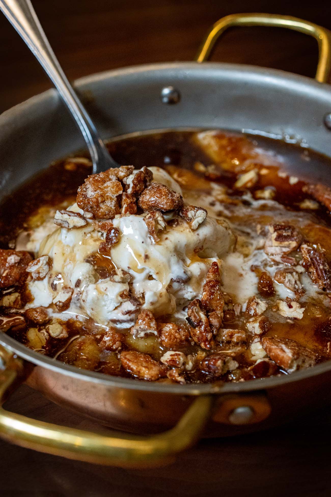 Bananas Foster served warm in a copper pan with brass handles, featuring caramelized bananas in a dark brown sugar sauce topped with a melting scoop of vanilla ice cream and scattered candied pecans. A spoon rests in the pan on a wooden table.