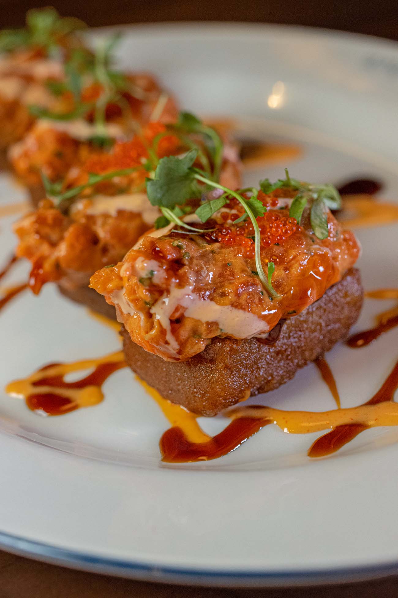  A close-up of the Spicy Salmon over Crispy Rice at WC Seafood Kitchen, showing individual bites of golden fried crispy rice topped with seasoned spicy salmon, bright orange tobiko, microgreens, and drizzles of aioli and eel sauce across a white plate.