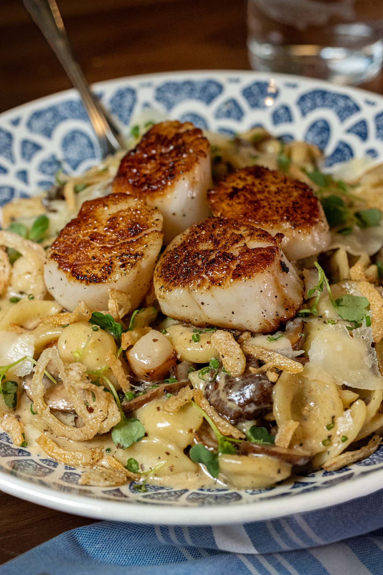 4 large seared scallops with deep golden-brown crusts resting on a bed of orecchiette pasta, mushrooms, crispy fried shallots, and microgreens in a garlic cream sauce, served in a blue and white patterned bowl with a fork resting at the edge.