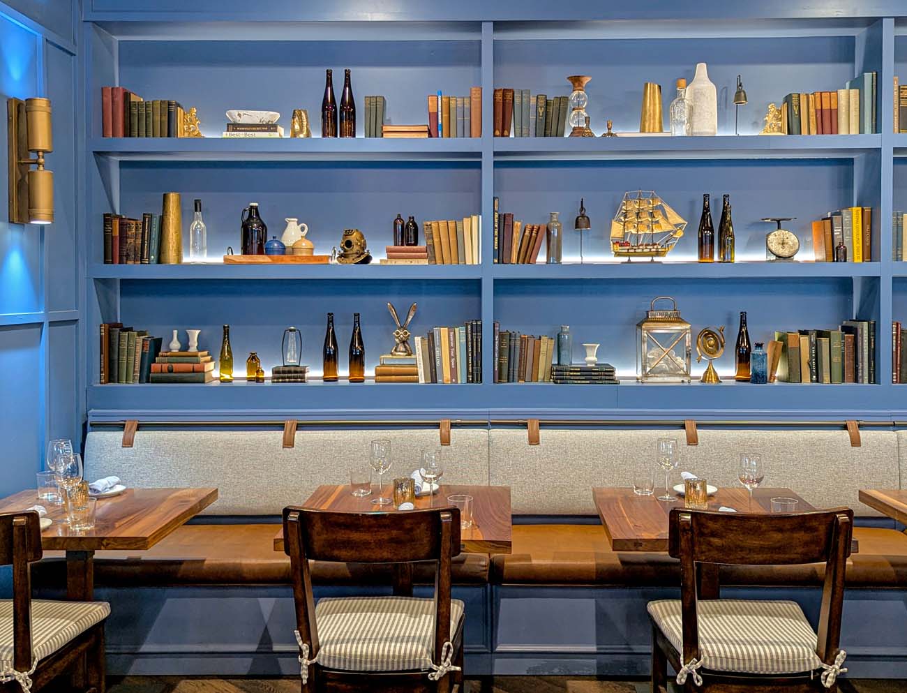  The dining room of WC Seafood Kitchen in West Chester, featuring a sweeping wall of blue built-in shelving filled with books, bottles, maritime curiosities including a vintage deep-sea diver's helmet, a model ship, and brass instruments. Wooden tables with striped upholstered chairs are set with wine glasses along a banquette, and brass wall sconces light the room.