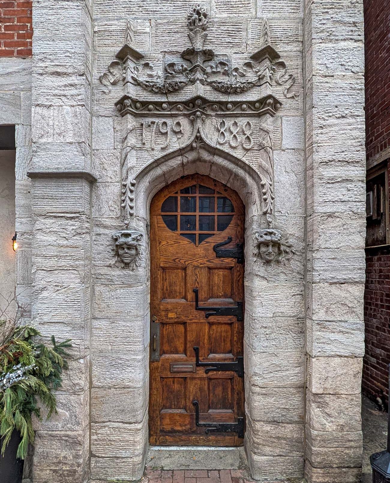 A Gothic-arched stone doorway in downtown West Chester, Pennsylvania, with ornate carved stonework, two carved face sculptures flanking the entrance, and the dates 1799 and 1888 carved above the wooden door with wrought iron hardware. A winter evergreen arrangement sits to the left.