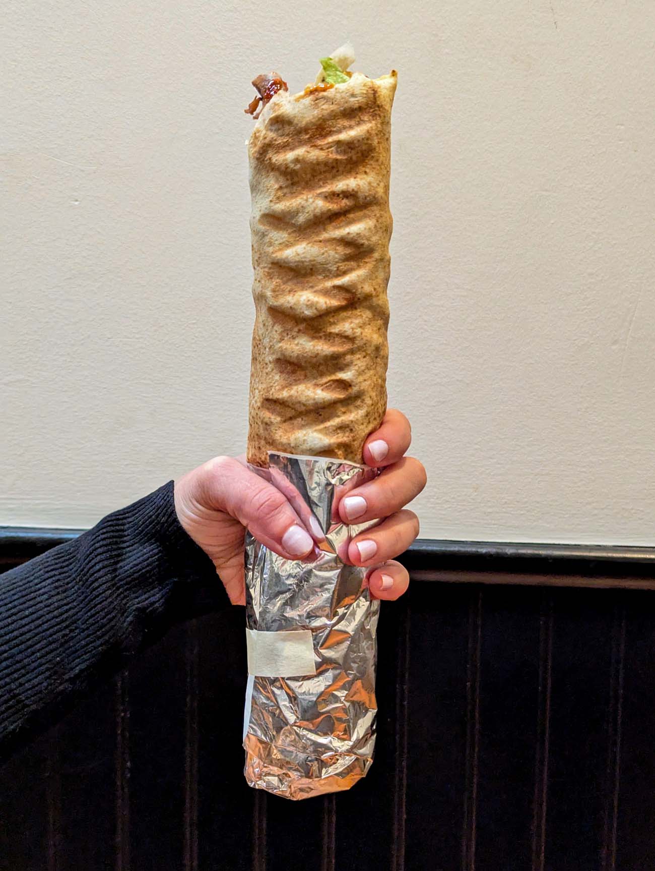 A hand holding a large foil-wrapped beef shawarma wrap from La Tartine in West Chester, with grilled pita visible and bits of lettuce and meat at the top. The background shows a dark wainscoted wall inside the casual cafe.