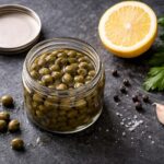 Small open glass jar of capers in brine, lid set to the side, a few loose capers scattered on the surface. Half a lemon, a sprig of fresh flat-leaf parsley, a few whole black peppercorns, a small clove of garlic.