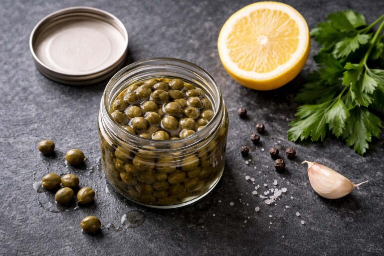 Small open glass jar of capers in brine, lid set to the side, a few loose capers scattered on the surface. Half a lemon, a sprig of fresh flat-leaf parsley, a few whole black peppercorns, a small clove of garlic.