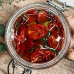 an open jar of sun-dried tomatoes on a wooden cutting board. herbs and ginger on the side.