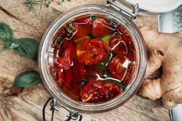 an open jar of sun dried tomatoes on a wood cutting board. herbs and ginger on the side.