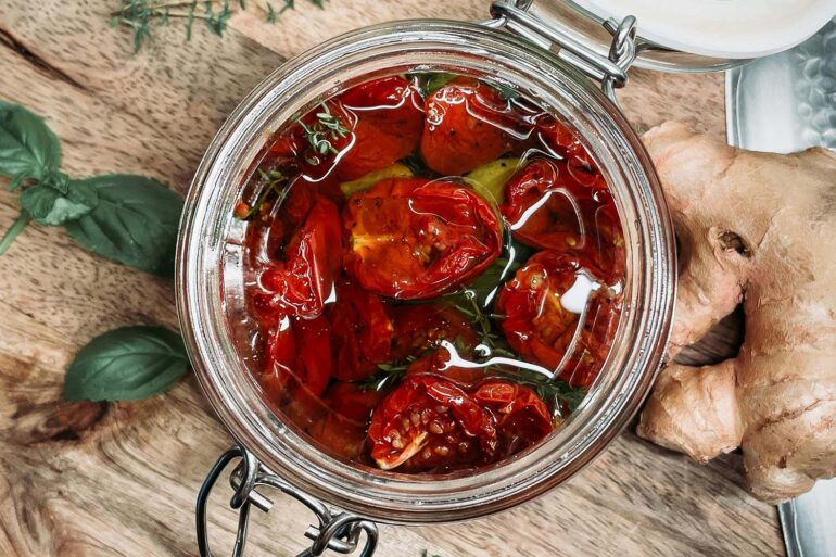 an open jar of sun dried tomatoes on a wood cutting board. herbs and ginger on the side.