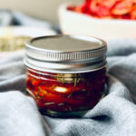 a jar of sealed sun dried tomatoes in a table with a dish towel around them. fresh tomatoes in the background.