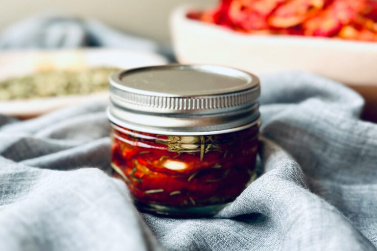 a jar of sealed sun dried tomatoes in a table with a dish towel around them. fresh tomatoes in the background.