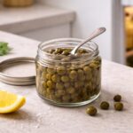 Open jar of capers in brine, lid beside it, a small spoon resting across the jar mouth. Refrigerator door slightly ajar in the right background, soft cold light spilling out. A lemon wedge and a few scattered capers in the foreground.