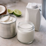 An open can of coconut milk with thick cream visible, a sealed carton of coconut milk beside it, a small glass container of poured coconut milk in front. Lime halves and a split fresh coconut in the background. Refrigerator door slightly ajar