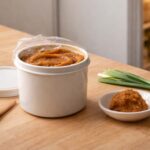 a small white ceramic dish with a tablespoon of miso paste scooped into it, a small bunch of trimmed green onions laid flat beside it, and a few sesame seeds scattered loosely on the counter. Far right, naturally soft in focus: the lower third of a matte white refrigerator door slightly ajar, warm interior light glowing, blurred shapes of other condiment containers visible on the door shelf.
