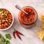 one open glass jar of red salsa with a spoon resting across the top. Left: a small white ceramic bowl of fresh pico de gallo with visible tomato, onion, and cilantro. Right: a loose handful of tortilla chips directly on the surface.
