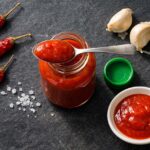 A small open glass bottle of red chili sauce, cap removed and set to the side, with a spoonful of bright red sauce resting across the mouth. Scattered around: three whole dried red chilies, two whole garlic cloves with papery skin slightly peeling, a small white ceramic dish with a pool of red sauce, a few grains of coarse sea salt.