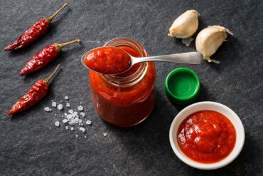 A small open glass bottle of red chili sauce, cap removed and set to the side, with a spoonful of bright red sauce resting across the mouth. Scattered around: three whole dried red chilies, two whole garlic cloves with papery skin slightly peeling, a small white ceramic dish with a pool of red sauce, a few grains of coarse sea salt.
