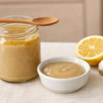 Tahini on kitchen counter. Fridge in the background. a pale cream linen countertop with soft natural light from above and slightly right. Left of frame an open glass jar of tahini with the lid resting beside it, pale golden sesame paste visible inside with a thin layer of oil sitting on top. A clean wooden spoon rests across the mouth of the jar suggesting it was just used.