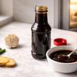 an open bottle of teriyaki sauce with cap resting beside it and a small white ceramic dish with a spoonful of sauce. Right side, the lower half of a white refrigerator door slightly ajar
