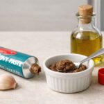 Pale stone counter. Open tube of anchovy paste with cap set beside it, a small ramekin with a teaspoon of paste, a clove of garlic, a small bottle of olive oil. Refrigerator door slightly ajar in the right background, soft cold light spilling out