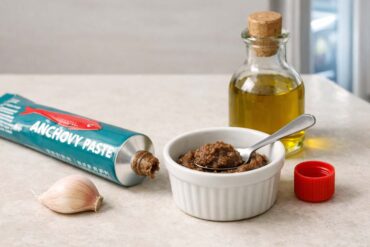 Pale stone counter. Open tube of anchovy paste with cap set beside it, a small ramekin with a teaspoon of paste, a clove of garlic, a small bottle of olive oil. Refrigerator door slightly ajar in the right background, soft cold light spilling out