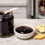 an open glass jar of hoisin sauce cap resting on its side next to the jar, dark glossy sauce visible inside. A small white ceramic spoon rests across the lip of the jar suggesting it was just used. Slightly right of center: a small round white ceramic dish with a tablespoon of hoisin sauce poured into it, a halved garlic clove cut-side up beside it showing the layered interior, and one thin diagonal slice of fresh ginger placed flat on the counter.