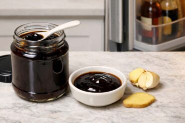 an open glass jar of hoisin sauce cap resting on its side next to the jar, dark glossy sauce visible inside. A small white ceramic spoon rests across the lip of the jar suggesting it was just used. Slightly right of center: a small round white ceramic dish with a tablespoon of hoisin sauce poured into it, a halved garlic clove cut-side up beside it showing the layered interior, and one thin diagonal slice of fresh ginger placed flat on the counter.