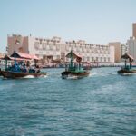 Traditional wooden abra water taxis carrying passengers across Dubai Creek, with UAE flags flying from the boats, historic low-rise heritage buildings along the Bur Dubai waterfront, and modern glass skyscrapers visible in the background under a clear blue sky.