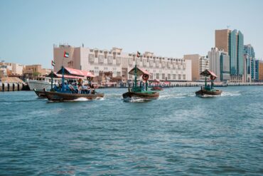 Traditional wooden abra water taxis carrying passengers across Dubai Creek, with UAE flags flying from the boats, historic low-rise heritage buildings along the Bur Dubai waterfront, and modern glass skyscrapers visible in the background under a clear blue sky.