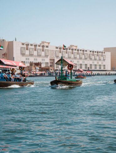 Traditional wooden abra water taxis carrying passengers across Dubai Creek, with UAE flags flying from the boats, historic low-rise heritage buildings along the Bur Dubai waterfront, and modern glass skyscrapers visible in the background under a clear blue sky.
