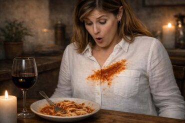 A woman with shoulder-length brown hair sits at a candlelit dinner table looking down at her chest with a shocked, open-mouthed expression. A large smear of tomato sauce covers the front of her white linen button-down shirt. In front of her on the table is a bowl of spaghetti with marinara sauce and a fork, a glass of red wine, and a lit white candle in the foreground. The background is a warmly lit, rustic stone interior.