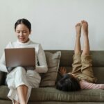 Cheerful mother working on laptop near daughter on couch