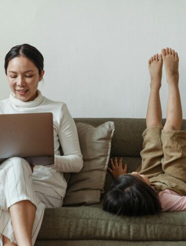 Cheerful mother working on laptop near daughter on couch