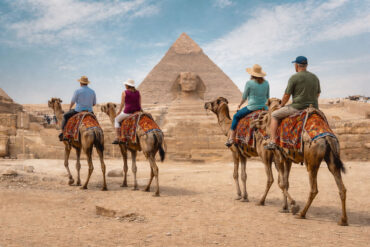 A wide, realistic desert scene at the Giza plateau shows the Great Sphinx of Giza centered in the midground with the Great Pyramid of Giza rising directly behind it under a bright, lightly clouded sky. In the foreground, four older tourists—two couples—ride separate camels spaced naturally apart as they travel toward the monuments. Each camel is standing and outfitted with a distinctly different, colorful woven saddle blanket featuring unique patterns and colors. The riders wear varied casual clothing, including sun hats, short-sleeve shirts, and light pants or shorts, with each person dressed differently to avoid repetition. The group is seen from behind, moving forward across sandy ground scattered with small rocks. Low stone ruins, pathways, and small clusters of distant visitors line the middle distance, reinforcing a busy but open archaeological setting.