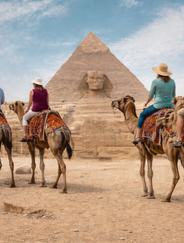 A wide, realistic desert scene at the Giza plateau shows the Great Sphinx of Giza centered in the midground with the Great Pyramid of Giza rising directly behind it under a bright, lightly clouded sky. In the foreground, four older tourists—two couples—ride separate camels spaced naturally apart as they travel toward the monuments. Each camel is standing and outfitted with a distinctly different, colorful woven saddle blanket featuring unique patterns and colors. The riders wear varied casual clothing, including sun hats, short-sleeve shirts, and light pants or shorts, with each person dressed differently to avoid repetition. The group is seen from behind, moving forward across sandy ground scattered with small rocks. Low stone ruins, pathways, and small clusters of distant visitors line the middle distance, reinforcing a busy but open archaeological setting.