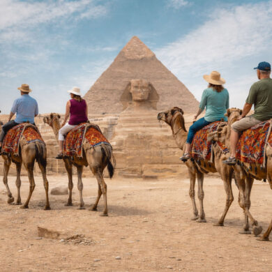 A wide, realistic desert scene at the Giza plateau shows the Great Sphinx of Giza centered in the midground with the Great Pyramid of Giza rising directly behind it under a bright, lightly clouded sky. In the foreground, four older tourists—two couples—ride separate camels spaced naturally apart as they travel toward the monuments. Each camel is standing and outfitted with a distinctly different, colorful woven saddle blanket featuring unique patterns and colors. The riders wear varied casual clothing, including sun hats, short-sleeve shirts, and light pants or shorts, with each person dressed differently to avoid repetition. The group is seen from behind, moving forward across sandy ground scattered with small rocks. Low stone ruins, pathways, and small clusters of distant visitors line the middle distance, reinforcing a busy but open archaeological setting.