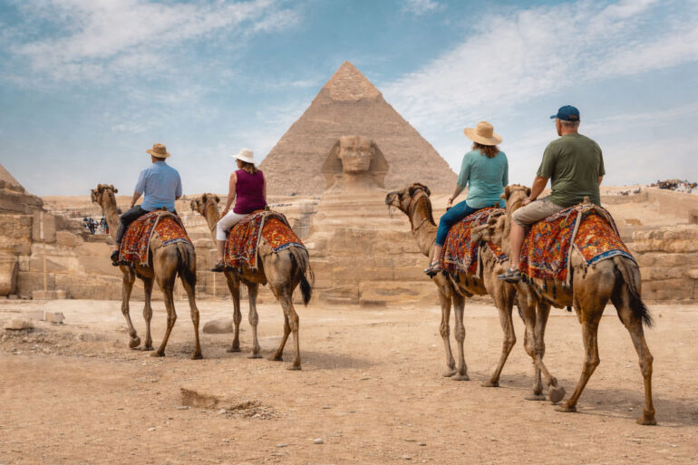 A wide, realistic desert scene at the Giza plateau shows the Great Sphinx of Giza centered in the midground with the Great Pyramid of Giza rising directly behind it under a bright, lightly clouded sky. In the foreground, four older tourists—two couples—ride separate camels spaced naturally apart as they travel toward the monuments. Each camel is standing and outfitted with a distinctly different, colorful woven saddle blanket featuring unique patterns and colors. The riders wear varied casual clothing, including sun hats, short-sleeve shirts, and light pants or shorts, with each person dressed differently to avoid repetition. The group is seen from behind, moving forward across sandy ground scattered with small rocks. Low stone ruins, pathways, and small clusters of distant visitors line the middle distance, reinforcing a busy but open archaeological setting.