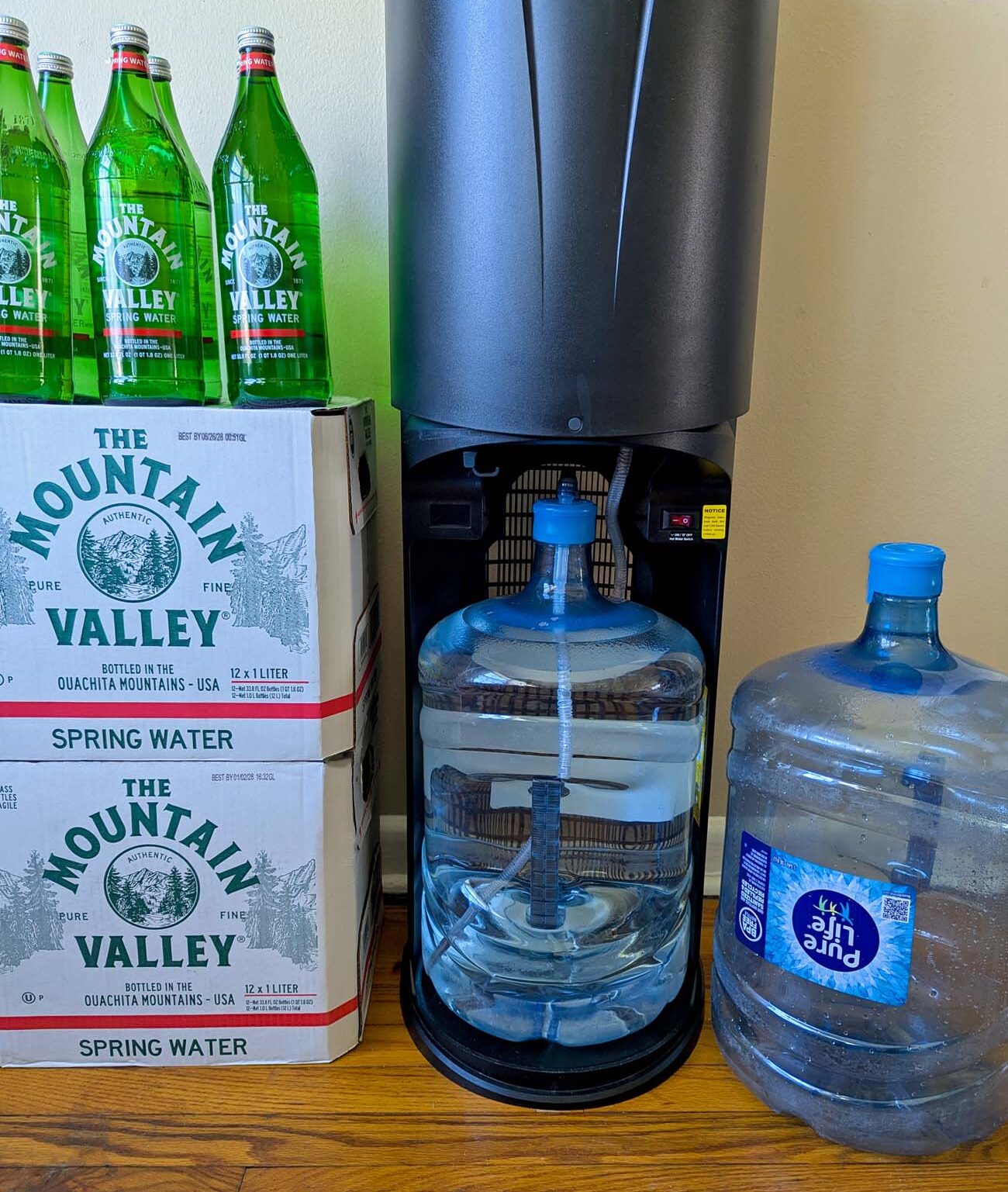 Primo bottom-load water dispenser with the cabinet door open showing a full five-gallon Pure Life jug loaded inside with the probe connected, flanked by two Mountain Valley cases and a spare Pure Life jug on the floor.
