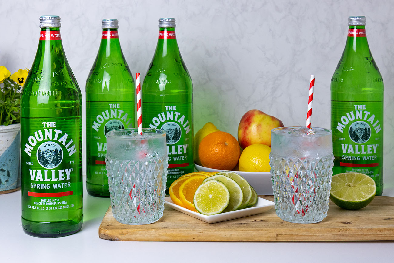 Four one-liter Mountain Valley spring water glass bottles arranged behind two crystal glasses with striped straws and sliced citrus on a wooden board, with fresh fruit in a bowl and yellow flowers in the background.