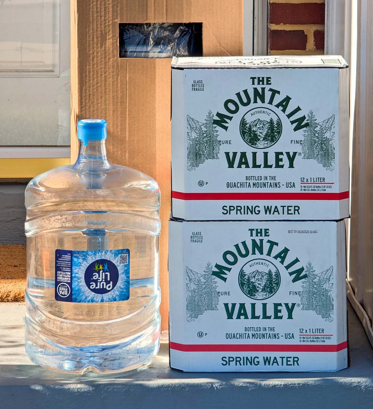 Vertical shot of a five-gallon Pure Life water jug beside two stacked Mountain Valley spring water cases on a front porch in golden morning light, with a brick wall and front door in the background.