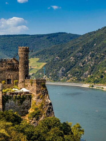 Burg Rheinstein castle perched on a rocky wooded cliff above the Rhine River in Germany, featuring medieval stone towers with battlements and arched Gothic windows, with terraced vineyard slopes and densely forested green hills rising on the opposite bank under a bright blue summer sky.