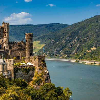 Burg Rheinstein castle perched on a rocky wooded cliff above the Rhine River in Germany, featuring medieval stone towers with battlements and arched Gothic windows, with terraced vineyard slopes and densely forested green hills rising on the opposite bank under a bright blue summer sky.