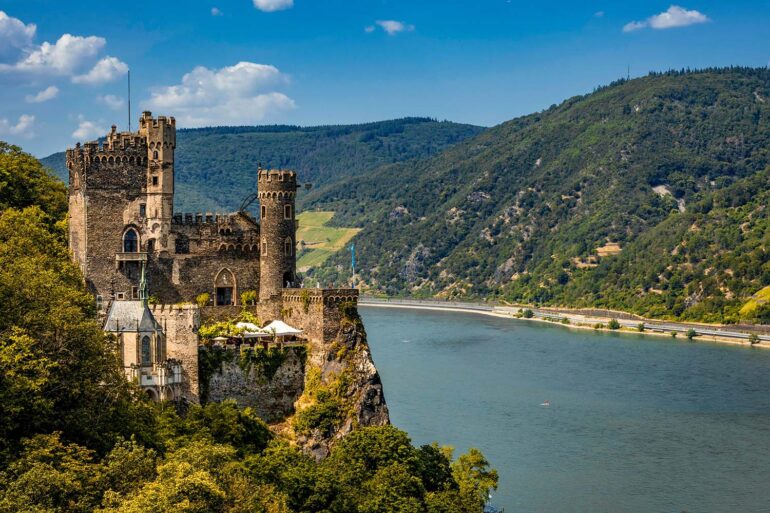 Burg Rheinstein castle perched on a rocky wooded cliff above the Rhine River in Germany, featuring medieval stone towers with battlements and arched Gothic windows, with terraced vineyard slopes and densely forested green hills rising on the opposite bank under a bright blue summer sky.
