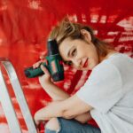 a woman with a drill on a ladder in front of a partially painted wall doing home maintenance
