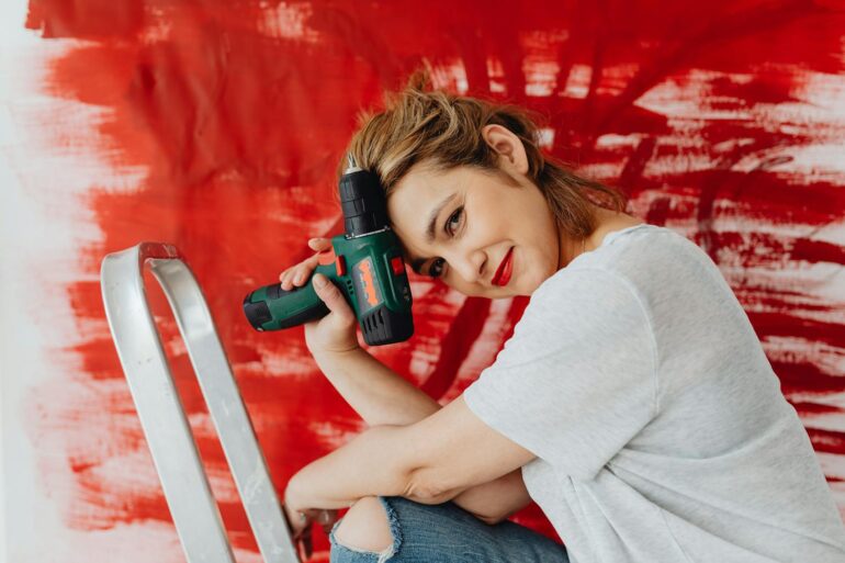 a woman with a drill on a ladder in front of a partially painted wall doing home maintenance