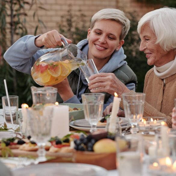 A younger woman smiling and pouring fruit-infused water for an older woman at a warmly lit outdoor dinner table with candles, fresh food, and family gathered around