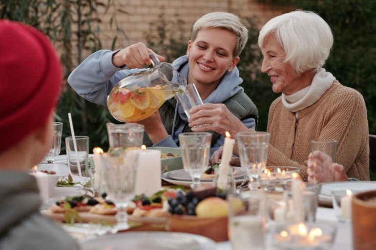 A younger woman smiling and pouring fruit-infused water for an older woman at a warmly lit outdoor dinner table with candles, fresh food, and family gathered around