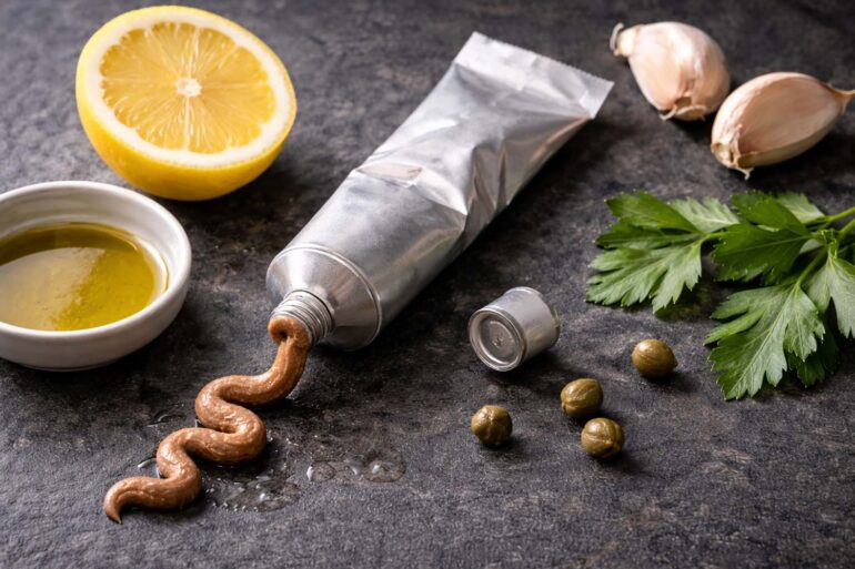 Dark slate surface. A small open tube of anchovy paste squeezed at one end, a few scattered capers, a halved lemon, two whole garlic cloves, a small dish of olive oil, a sprig of fresh flat-leaf parsley.