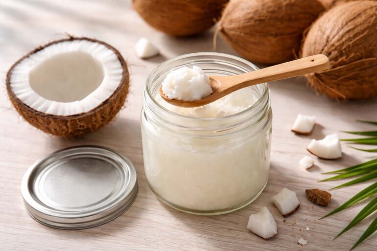 An open glass jar of white solid coconut oil, lid resting beside it. A clean wooden spoon with a small scoop of coconut oil resting across the jar mouth. Scattered around the jar: a halved fresh coconut