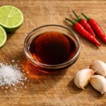 A top-down flat lay of a small glass bowl of dark amber fish sauce on a worn wooden cutting board. Surrounding the bowl: two halved limes showing bright green flesh, three fresh red Thai chili peppers, a small pile of coarse sea salt crystals, and three whole garlic cloves with papery skin. Natural side window light casting soft shadows to the right. Muted, warm tones.