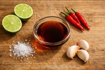 A top-down flat lay of a small glass bowl of dark amber fish sauce on a worn wooden cutting board. Surrounding the bowl: two halved limes showing bright green flesh, three fresh red Thai chili peppers, a small pile of coarse sea salt crystals, and three whole garlic cloves with papery skin. Natural side window light casting soft shadows to the right. Muted, warm tones.