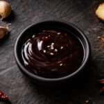 a small round matte black ceramic dish filled with thick, dark brown glossy hoisin sauce, sesame seeds scattered naturally on the surface of the sauce. Surrounding the dish whole garlic cloves with papery skin slightly peeling at the edges, one whole star anise, a thin diagonal slice of fresh ginger root showing pale yellow flesh at the cut edge, a small loose pinch of Chinese five-spice powder spilled directly onto the slate, and two small dried red chilies